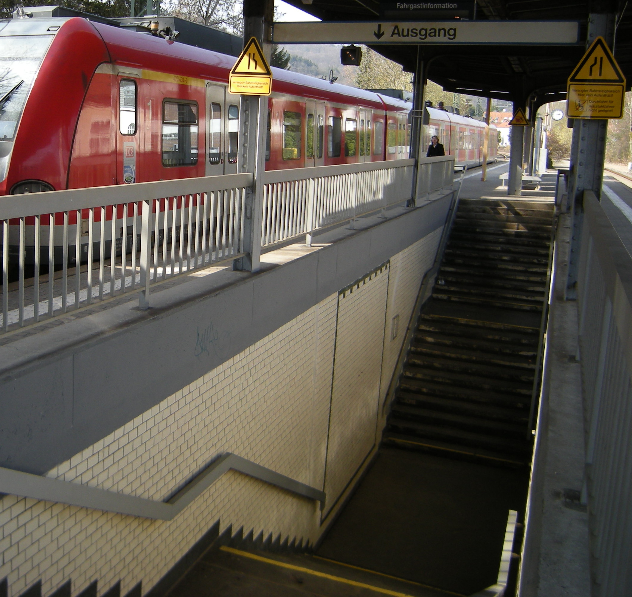 Zu sehen sind die Zugangstreppen zum Bahnhof Lorsbach, Teile des Bahnsteigs und eine S-Bahn in Richtung Frankfurt. Es fehlt ein barrierefreier Zugang zum Mittelsteig.