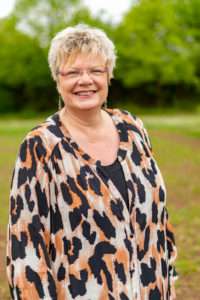 Karin Lübbers ist circa 50 Jahre alt, die trägt kurze blonde Haare, eine Brille und lange Ohrringe. Sie trägt ein Kleid mit Leopardenmuster. Sie steht auf einer Wiese mit Bäumen im Hintergrund.