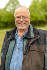 Das Bild zeigt Michael Mehr. Michael ist circa 60 Jahre alt. Er hat kurze graue Haare und einen grauen Vollbart. Er trägt eine Brille. Er hat eine braune Jacke an und steht auf einer Wiese vor Bäumen.