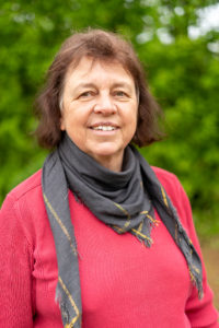 Das Bild zeigt Barbara Grassel. Barbara ist Mitte sechzig, hat braune schulterlange Haare. Sie steht auf einer Wiese vor Bäumen und trägt ein graues Halstuch und einen roten Pullover.