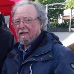 Das Bild zeigt Heinz Michael Merkel. Heinz ist circa 70 Jahre alt, er hat graue Haare und einen grauen Vollbart. Er trägt eine Brille, eine blaue Jacke und steht vor einem roten Zelt.