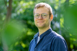 Konstantin ist Anfang Dreißig, hat kurze braune Haare, trägt eine Brille und Bart. Auf dem Bild in der Natur trägt ein blaues Hemd mit offenem Kragen