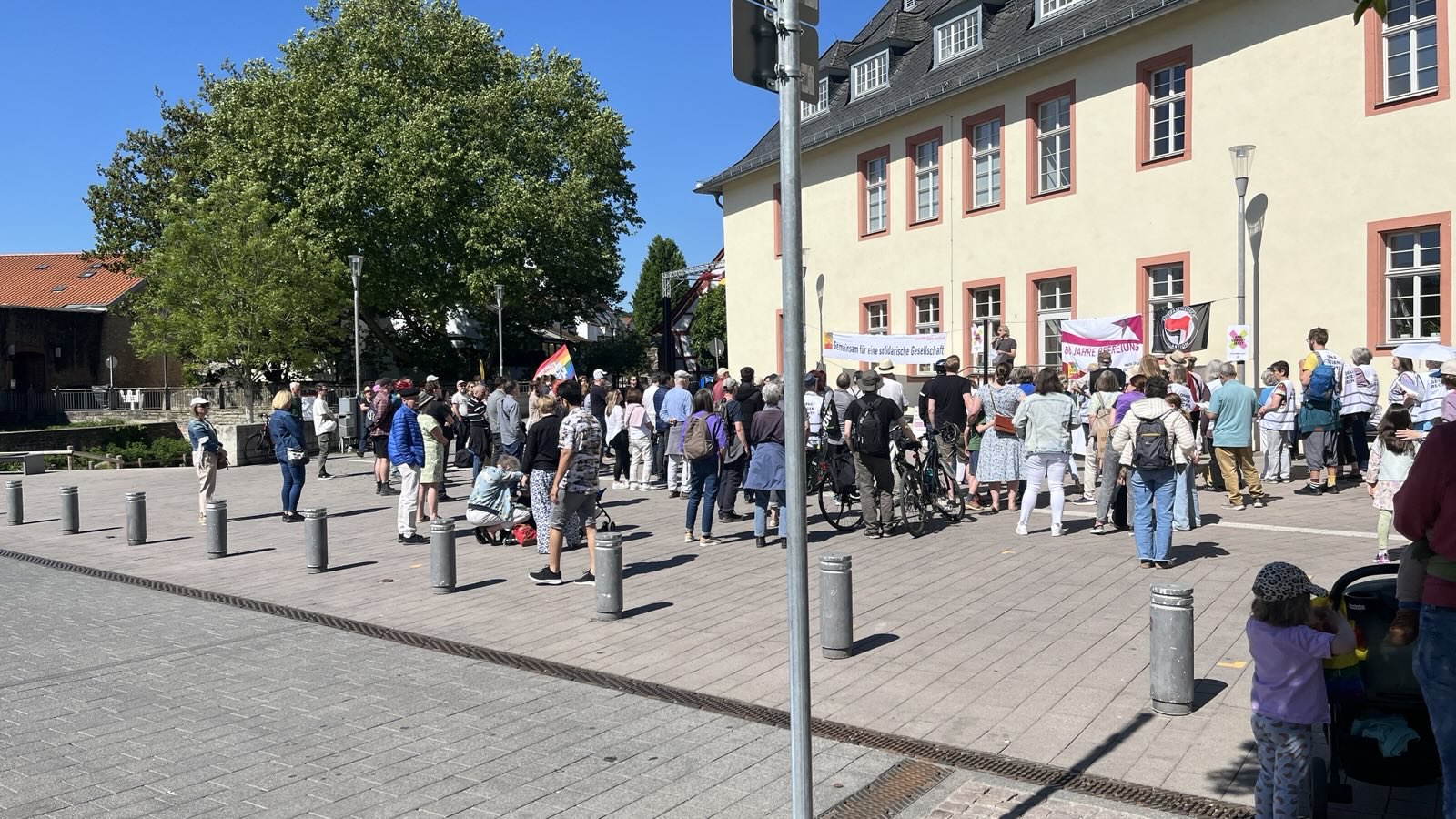 Viele Menschen stehen auf einem Platz vor einem gelben Gebäude im schönsten Sonnenschein