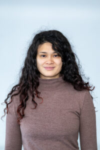 Eine Frau Mitte 20 mit langen schwarzen Locken in einem braunen Pullover