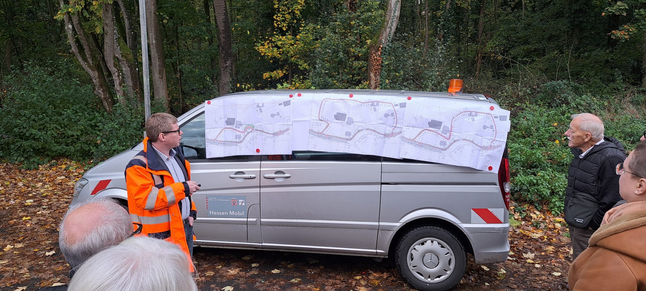An einem Kleinbus sind verschiedene Routenplanungen für den Radweg zwischen Lorsbach und Hofheim angepinnt. Eine Gruppe von Menschen betrachtet die Entwürfe.