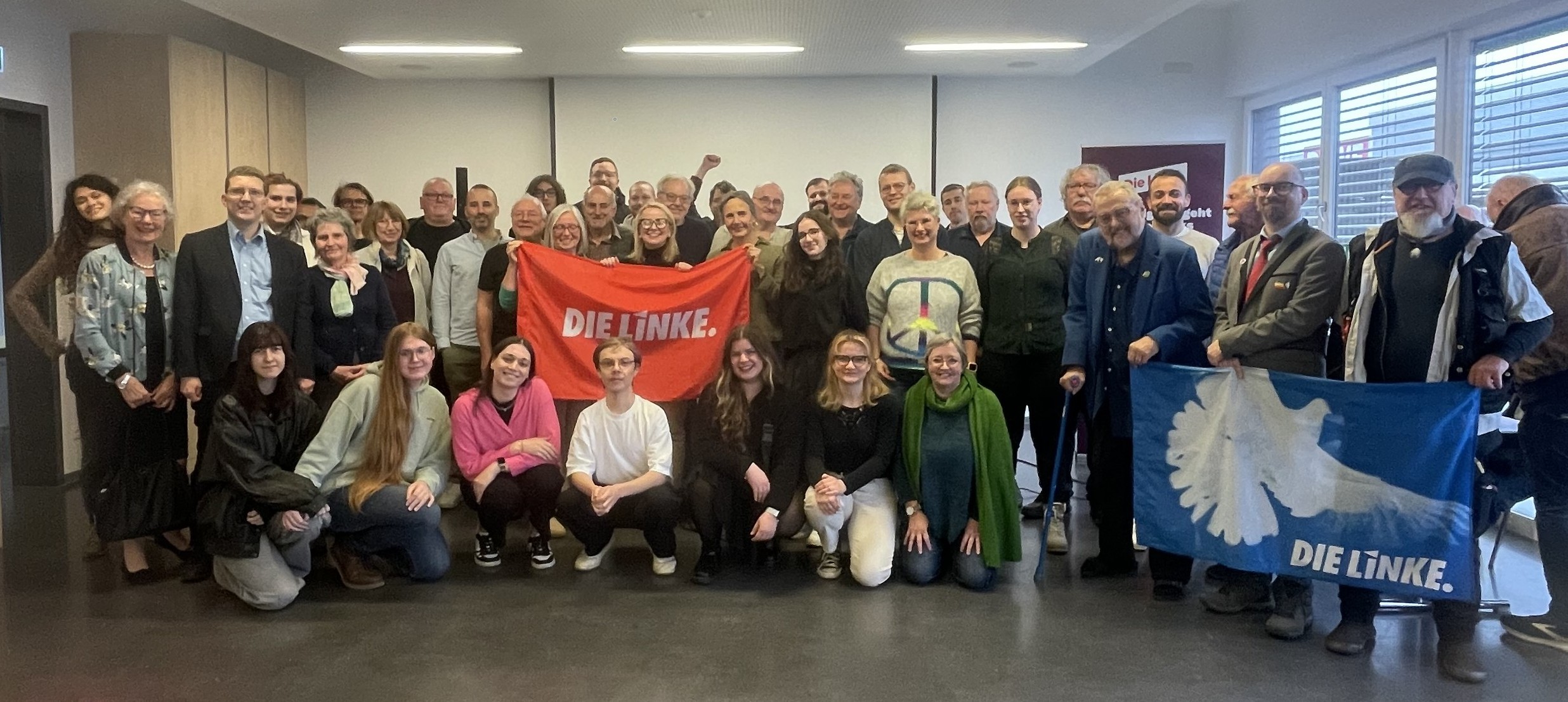 Eine bunte Gruppe von ca. 40 Personen. Sie halten eine Friedensfahne und eine Fahne der Partei Die Linke hoch.