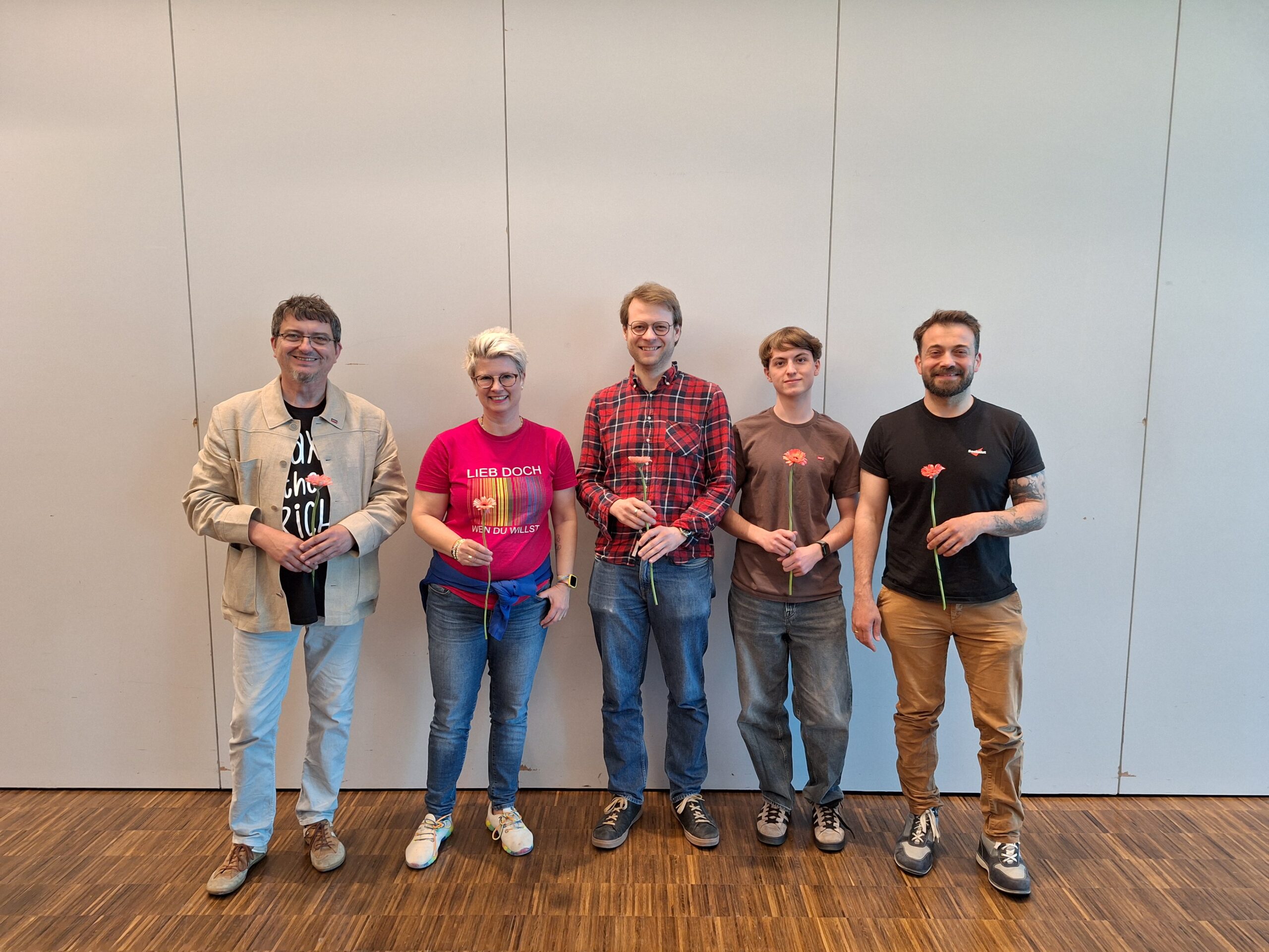Fünf Personen stehen vor einer weißen Wand und halten Blumen in der Hand