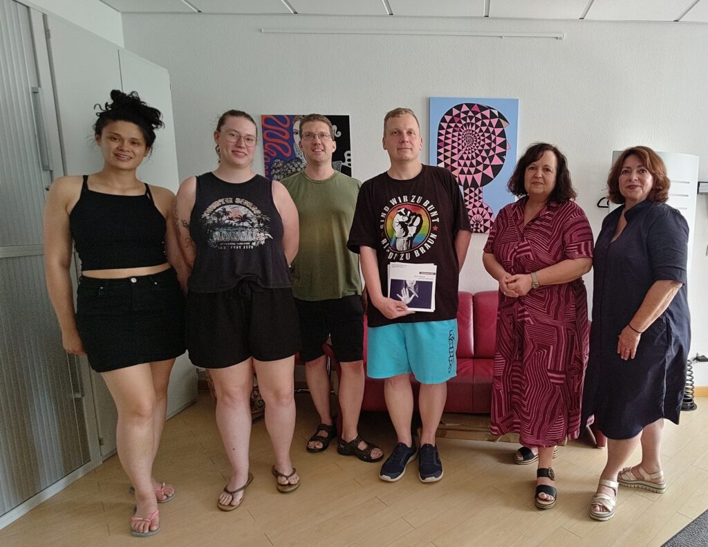 In den Geschäftsräumen von Frauen helfen Frauen Main-Taunus stehen vkier Frauen und zwei Männer für ein Gruppenbild vor zwei schönen Kunstwerken.